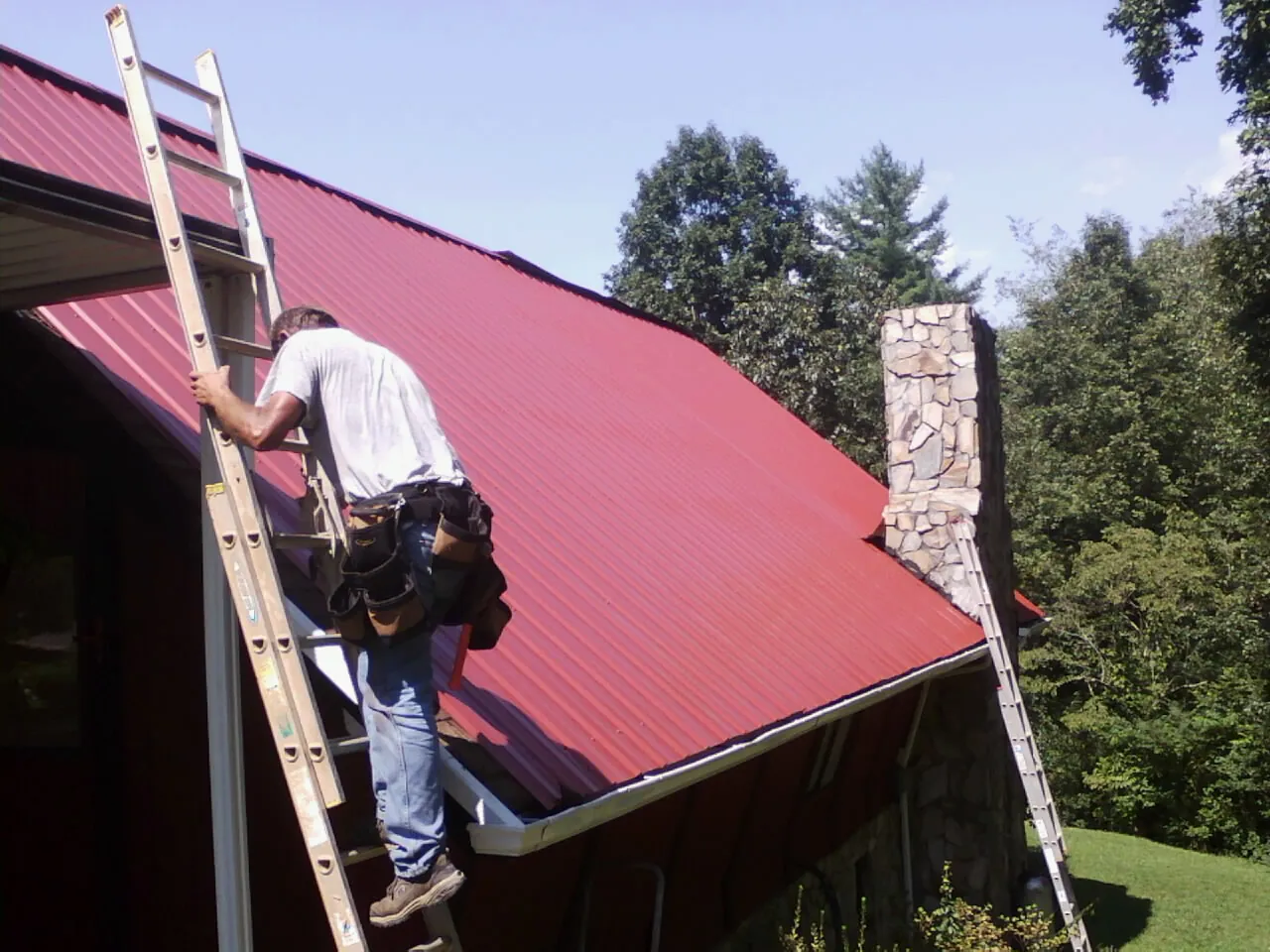 Roofer on a ladder beside a red metal roof and stone chimney — Soffit & Fascia Repair by Roofing Brothers in Shamokin